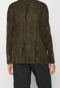 Olivgrön stickad tröja med lös passform, med slitna detaljer, långa ärmar och en texturerad design. Mönster av revor finns genomgående.