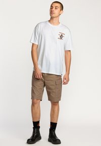 T-shirt blanc à manches courtes avec un logo graphique orange sur la poitrine, associé à un short cargo beige avec des poches latérales et des bottes noires à la cheville.