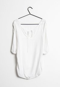 Blouse blanche à coupe ample, manches courtes, encolure profonde en arrondi, avec des liens à l'avant. Présente des détails texturés sur les coutures des épaules.