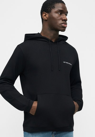 Άνδρας που φοράει μαύρο φούτερ με κουκούλα και μπροστινή τσέπη, με μικρό λευκό κείμενο "LES BENJAMINS" στο στήθος, τα χέρια στις τσέπες, κοιτάζει προς το πλάι.