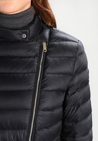 Svart dunjacka med horisontellt quiltat mönster, hög krage och en silverfärgad dragkedja. Slät textur med subtil glans.