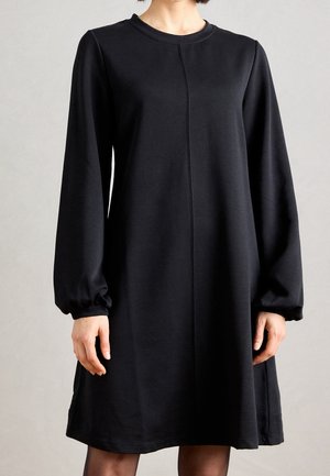 Femme portant une robe noire simple à manches longues avec un col rond et des poignets légèrement bouffants, debout devant un fond uni.