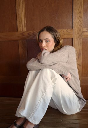 Beżowy sweter z dzianiny, luźne białe spodnie i ciemne buty, siedząca na tle drewnianej ściany z poziomymi panelami, z nogami przytulonymi do ciała.