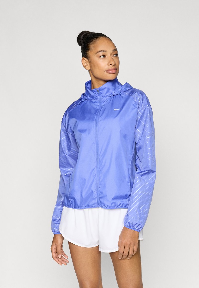 Ελαφρύ μωβ αδιάβροχο μπουφάν με κουκούλα, φερμουάρ μπροστά και μανίκια με διάτρητο δίχτυ. Φορεμένο πάνω από λευκά σορτς, με ελαστικές μανσέτες.