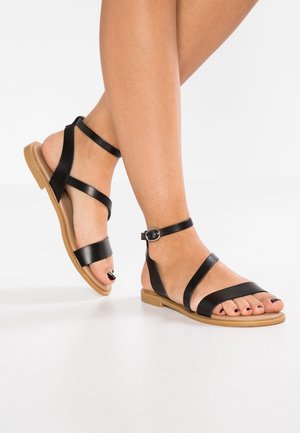 Füße, die schwarze flache Ledersandalen mit Knöchelriemen und hellbrauter Sohle auf weißem Hintergrund tragen.