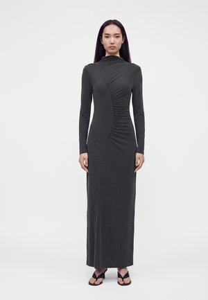 Femme aux longs cheveux raides portant une robe longue moulante gris foncé à manches longues avec des détails froncés et des sandales noires à talons.