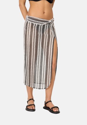 Mujer con top de bikini negro y falda envolvente a rayas con abertura lateral, combinada con sandalias negras planas con tiras.