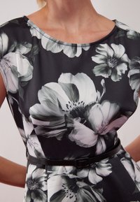 Robe noire à fleurs comportant de grandes fleurs blanches et grises, manches courtes, col rond et une ceinture noire ajustée à la taille.