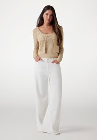 Beige knitted cardigan with open front and geometric pattern, paired with wide-leg white trousers. Smooth texture, relaxed fit, minimal hardware.
