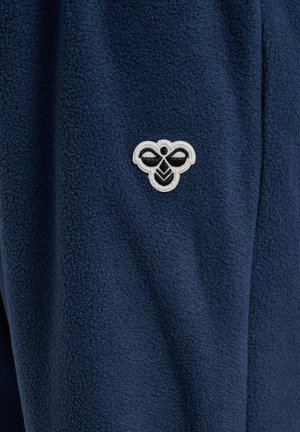 Teksturowana tkanina granatowa z małym białym i czarnym haftowanym logo pszczoły blisko środka.