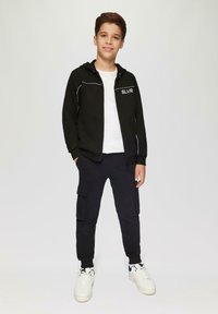 Zwarte rits-hoodie met witte accenten en het "SLVR" logo over een wit shirt, gecombineerd met zwarte cargobroeken en witte sneakers.