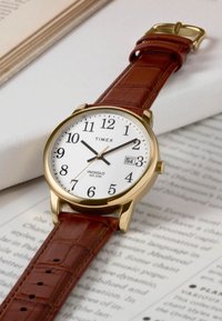 Montre Timex avec un boîtier doré, un cadran blanc, des chiffres arabes noirs et un bracelet en cuir brun à motif crocodile. Équipée d'une fonction date.