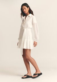 Bílá minišaty z očka s dlouhými rukávy, zapínáním na knoflíky u límce, řaseným pasem a rozšířenou sukní. Nošeno s černými žabkami.