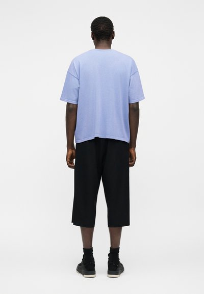 Άνδρας που στέκεται γυρισμένος με την πλάτη φοράει ανοιχτό μπλε κοντομάνικο πουκάμισο, μαύρες βερμούδες μέχρι το γόνατο, μαύρες κάλτσες και μαύρα αθλητικά παπούτσια.