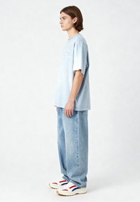 T-shirt oversized blu chiaro con stampa grafica bianca, abbinata a jeans blu chiaro a vestibilità ampia e sneakers bianche con accenti rossi e blu.