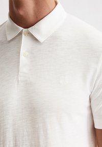 Witte poloshirt gemaakt van een lichte, gestructureerde stof met een kraag, twee knopen en een geborduurd logo aan de linkerkant.