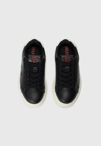 Une paire de baskets noires avec des semelles blanches, des lacets noirs et le texte rouge "HUGO YOUR WAY" sur la languette, vue de dessus sur fond blanc.