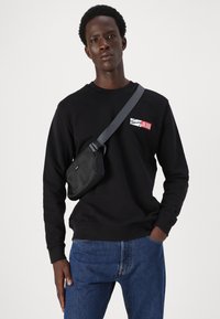 Svart genser med ribbestrikkede mansjetter, som har en fargeblokk-logo i rødt og hvitt. Kombinert med blå denimjeans og en svart crossover-bag.