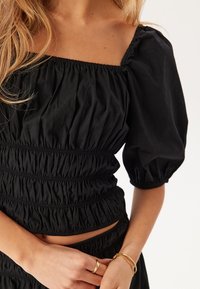 Top nero a maniche puff con scollatura quadrata, arricciatura elastica in vita e tessuto morbido e testurizzato. Accompagnato da semplice gioielleria dorata.