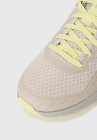 Lys grå sportsko med mesh-overdel, gule lisser og teksturert såle med detaljer og utskjæringer. Nærbilde av tåområdet.