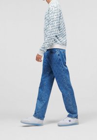 Felpa bianca con il logo blu "KARL LAGERFELD" ripetuto, abbinata a pantaloni in denim blu con un motivo testurizzato e scarpe da ginnastica bianche.