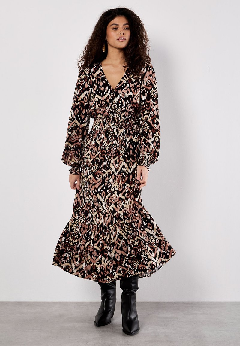 Maxikleid mit einem abstrakten Muster in Schwarz, Pink und Creme, langen Puffärmeln, tailliertem Bund und einem volantrimierten Saum. Kombiniert mit schwarzen Stiefeln.
