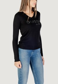 Maglione nero a maniche lunghe con scollatura a V e dettagli in strass logo, abbinato a jeans azzurri chiari, che mettono in risalto una silhouette aderente.