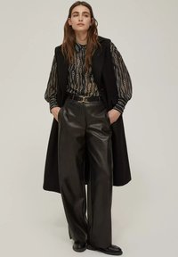 Pantaloni neri in pelle a gamba larga abbinati a una camicetta a fantasia e un gilet nero, con una cintura con fibbia circolare.