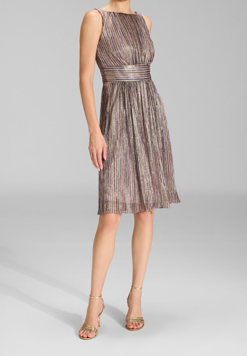 Femme portant une robe sans manches mi-longue aux rayures verticales métalliques multicolores et des talons hauts dorés à brides assortis sur fond gris uni.