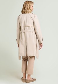 Mujer rubia vista desde atrás, vestida con un abrigo trench beige con cinturón, pantalones beige recortados y mocasines beige sobre un fondo pálido.