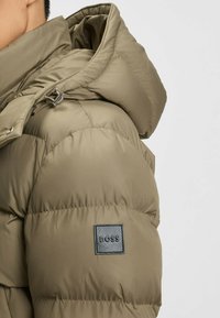 Bomber-lignende pufferjakke i olivengrøn med quiltet tekstur. Har en hætte og en rektangulær sort læderlogo-patch.