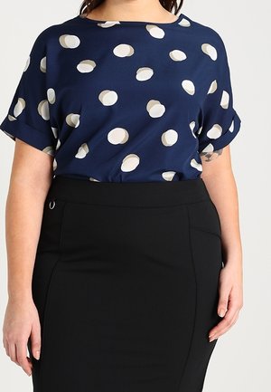 Mujer con una blusa de manga corta azul marino con lunares blancos metida dentro de una falda lápiz negra de cintura alta.