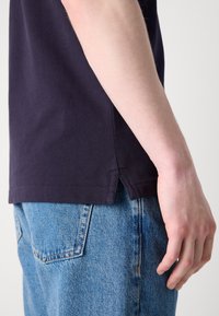 Personne portant une chemise foncée et un jean bleu, montrant la couture latérale et les détails de l'ourlet de la chemise près de la taille, la main détendue le long du corps.
