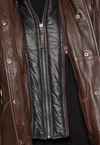 Brun läderjacka med ett avtagbart svart kviltat foder. Har dragkedjor och tryckknappar, vilket framhäver kontrasterande texturer och material.