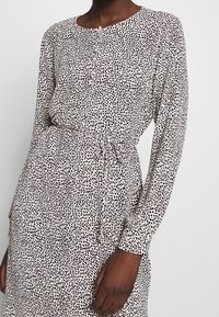 Dlouhé šaty s kulatým výstřihem, zdobené černo-bílým puntíkovaným vzorem. Obsahují zavazování v pase a zapínání na knoflíky.