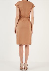 Lyhyt ruskea mekko, jossa on lyhyet hihat, joustava vyötärö ja polveen ulottuva helma. Pukeutunut vaaleanruskeisiin korkokenkiin.