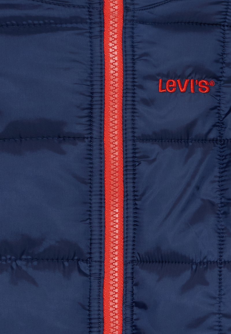 Chaqueta acolchada azul marino con un brillante cierre rojo y logo bordado. Material suave y brillante con textura acolchada y costuras visibles.