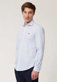 Camisa de manga larga a rayas blancas y azul claro con cuello abotonado y logo en acento azul marino. Tejido de algodón, corte ajustado.