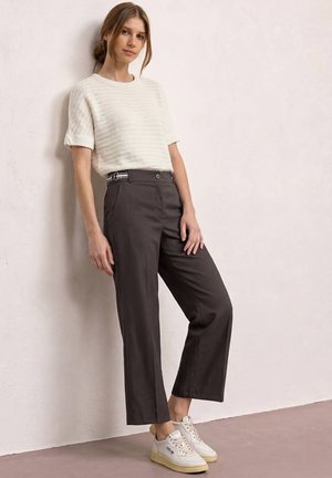 Jonge vrouw die leunt tegen een witte, gestructureerde muur, gekleed in een witte korte mouw trui, donkere cropped broek en witte sneakers.