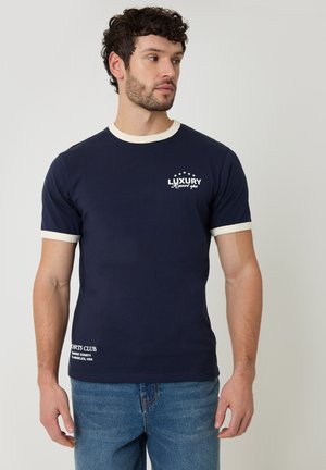 Uomo che indossa una T-shirt a maniche corte blu navy con rifiniture bianche e testo stampato, abbinata a jeans blu, che guarda verso sinistra.