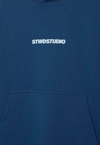 Sininen kangashuppari, jossa on etutasku ja valkoinen "STWDSTUDIO" -tekstilogo keskitettynä taskun ylle.