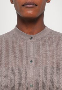 Pletený cardigan v svetlohnedej farbe s textúrovaným vzorom. Disponuje vysokým golierom a piatimi gombíkmi, vyrobený z mäkkej, jemne priesvitnej tkaniny.