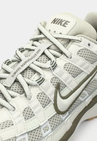 Γκρι και λευκό αθλητικό παπούτσι Nike με διάτρητο επάνω μέρος, υφασματικές λεπτομέρειες και χοντρές κορδόνια. Διαθέτει εμφανές λογότυπο Swoosh και μοναδικό μοτίβο πλέγματος.