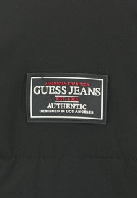 Sort stofmærke med hvid og rød tekst; indeholder "GUESS JEANS," "EST. 1981," "AUTENTISK," og "DESIGNET I LOS ANGELES."