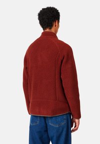 Giacca in pile rosso con colletto alto e superficie texture. Ha una vestibilità rilassata ed è abbinata a jeans di denim blu.