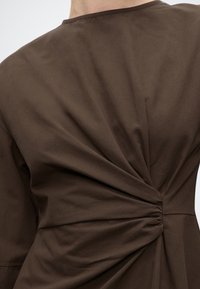 Κοντινή λήψη καφέ ρούχου κορυφής με σχεδιαστική λεπτομέρεια στριφτού, μαζεμένου υφάσματος στην πλάτη κοντά στη μέση.