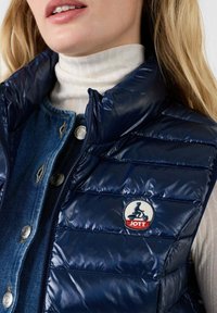 Nahaufnahme einer Frau in einem weißen Rollkragenpullover, einer blauen Jeansjacke und einer marineblauen gesteppte Weste mit einem JOTT-Logo-Patch auf der Brust.