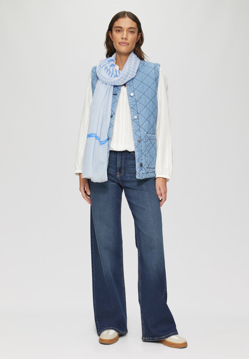 Gequilted blauwe vest met knoopsluiting, gecombineerd met een lichtblauwe gestreepte sjaal, witte blouse en snel brede donkere jeans. Casual schoenen inbegrepen.