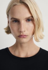 Černé triko s kulatým výstřihem, nošené osobou s krátkými, světlými blond vlasy a modrýma očima, na neutrálním pozadí.