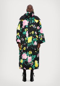 Abrigo acolchado de tela negra con gran estampado floral en rosa y amarillo. Cuenta con cuello alto, mangas largas y dos bolsillos frontales.